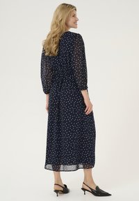 Mörkblå klänning med vita prickar, genomskinliga långärmade ärmar, insydd midja och flödande kjol; kombinerad med svarta slingbacks.