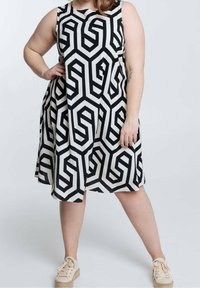 Persoon met een mouwloze, knielange jurk met zwart-wit geometrisch patroon en beige plateau sneakers, staand met de hand op de heup.