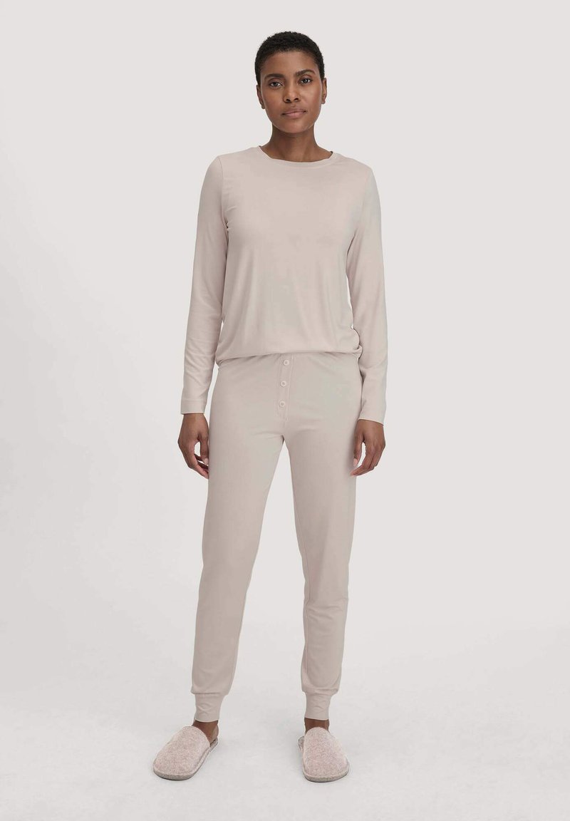 Helles beiges Loungewear-Set aus weichem Material, bestehend aus einem Langarm-Top und einer Hose mit elastischem Bund und Knopfdetails, kombiniert mit kuscheligen Hausschuhen.