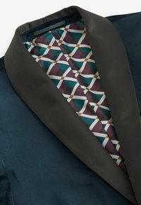Sötétkék bársony blézer fekete selyem stóla gallérral; a bélés geometrikus minta teal, burgundi és fehér színekben.