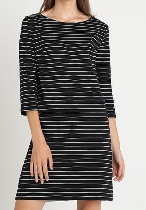 Femme portant une robe noire avec des rayures blanches horizontales et des manches trois-quarts, debout devant un fond clair uni.