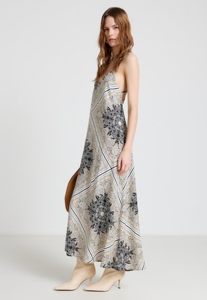 Femme portant une robe maxi sans manches à motifs et des talons beige à bout pointu, tenant un sac à main rond marron sur un fond uni.