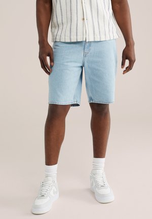 Mand iført lyseblå denimshorts, hvide sneakers, hvide sokker og en hvid kortærmet skjorte med vertikale striber.