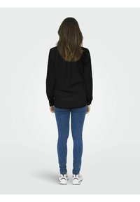 Chemise noire à manches longues avec poignets boutonnés, assortie à un jean bleu coupe slim et des baskets blanches. Les cheveux tombent en vagues lâches au-delà des épaules.