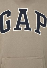 Tissu beige avec de grandes lettres noires bordées de blanc formant le mot "GAP" au-dessus d'une couture de poche horizontale.
