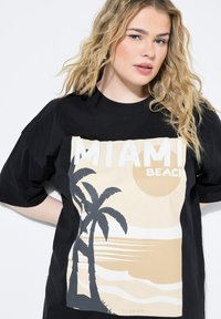 Musta puuvillapaita, jossa on kerma värinen grafiikka palmupuista, auringosta ja teksti "MIAMI BEACH" lihavoituna. Paidassa on rento malli ja lyhyet hihat.