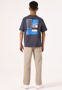 Camiseta de algodón gris con un gráfico geométrico en azul y blanco en la parte posterior, combinada con pantalones rectos beige y zapatos blancos.