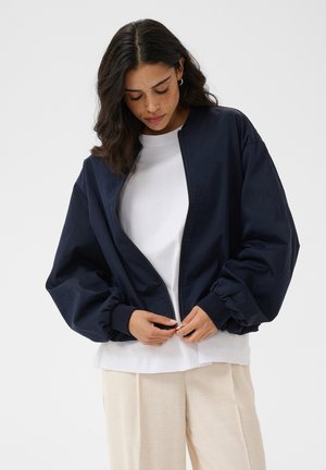 Femme aux cheveux foncés en train de dézipper une veste oversize bleu marine portée sur une chemise blanche et un pantalon beige sur un fond uni.