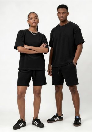 Dwie osoby stojące obok siebie, ubrane w czarne koszulki, czarne szorty i pasujące czarne buty sportowe Adidas na białym tle.