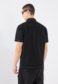 Camicetta nera a maniche corte con orlo dritto, cuciture a contrasto bianche e colletto classico. Indossata con pantaloni neri strappati.