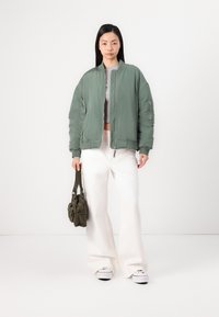 Veste bomber verte avec fermeture éclair, poignets côtelés et poches latérales, associée à un pantalon large blanc et un sac à main vert texturé.
