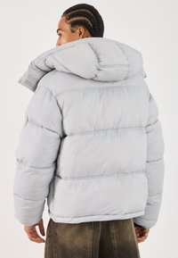 Siva puffer jakna s visokim ovratnikom, oversized dizajn, kližasti tekstura i vidljivi detalji šivanja. Uključuje kapu.