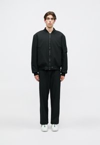 Melna bomber jaka ar rāvējslēdzēju priekšpusē, ribotiem piedurknēm un sānu kabatu, kombinēta ar melniem biksēm un baltām sporta apaviem. Vienkrāsaina, minimāla dizaina.