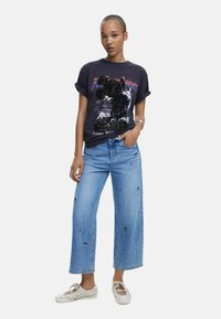 Dunkles Grafik-T-Shirt mit umgeschlagenen Ärmeln, hellblaue weite Jeans mit bestickten Patches und weiße Sneakers mit Riemendetails.