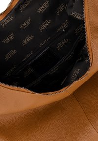 Innenraum einer tanfarbenen Ledertasche mit schwarzem Stofffutter, das mit wiederholtem, goldfarbenem "Venezia"-Branding versehen ist, sowie einem Reißverschlusspunkt für die Aufbewahrung.