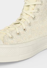 Κοντινό πλάνο ενός κρεμ sneaker ψηλού τύπου με λουλουδάτα δαντελωτά σχέδια, λαστιχένια σόλα και κορδόνια σε αντίστοιχο κρεμ χρώμα διαπερασμένα μέσα από μεταλλικά ρούχα.
