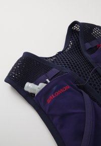 Purple hydration vest with mesh back, adjustable straps, and a pocket containing a water bottle. Salomon logo in red on the front.