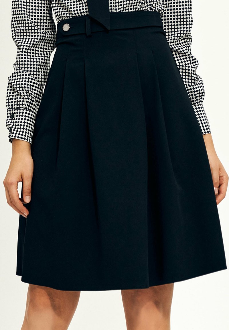 Kobieta w czarną plisowaną spódnicę o wysokim stanie oraz czarno-białą koszulę w kratę z długim rękawem, włożoną w spódnicę.