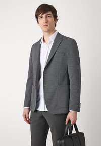 Blazer grigio strutturato con design a un petto, due tasche frontali, abbinato a una camicia bianca e pantaloni grigi, mentre tiene una borsa nera.