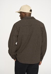 Brun ternet skjorte med l�øs pasform, der har krave, lange ærmer og et tekstureret stof. Kombineret med en lysefarvet kasket.