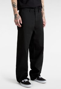 Schwarze, weit geschnittene Hose aus Baumwolle mit glatter Textur. Verfügt über Vordertaschen und einen klassischen Bund. Getragen mit schwarzen Turnschuhen.