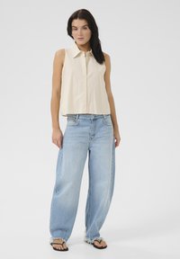 Krémová bezrukávová košeľa s gombíkmi skombinovaná so svetlo modrými oversize džínsami s voľným strihom a roztrhnutým okrajom, obutá s čiernymi flip-flopami.