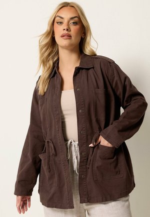 Blonde woman wearing a dark brown buttoned jacket with large pockets and a beige top, standing against a plain light background.