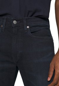 Personne portant un jean bleu foncé et une chemise bleu marine, debout avec un bras détendu sur le côté, vue de la taille jusqu'à mi-cuisse.