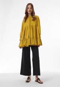 Blusa mostaza amarilla de volantes con cuello y mangas largas, combinada con pantalones negros de pierna ancha y sandalias planas negras con un acento dorado.