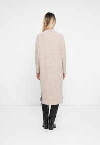 Lunga cardigan beige in maglia con collo alto, polsini a costine e spacco laterale, indossata sopra stivali neri con il tacco alto. Design minimalista, texture liscia.
