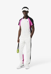 Tenue de sport comprenant un polo blanc avec des panneaux roses et noirs, un pantalon blanc avec un accent rose sur le côté et des chaussures blanches avec une marque.