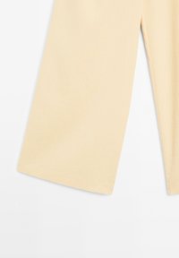 Gros plan d'un pantalon beige clair à jambes larges montrant des ourlets droits et une texture de tissu lisse sur fond blanc uni.