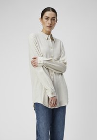Chemise beige clair à boutons en tissu texturé, avec un col et des manches longues, assortie à un pantalon en denim bleu.