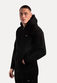 Sudadera con cremallera negra hecha de tela suave, con un bolsillo frontal, capucha ajustable y puños de canalé. Diseño minimalista con una pequeña etiqueta de logo.
