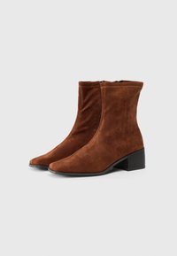 Botine din suede maro cu toc chunky și fermoare laterale, pe un fundal simplu deschis.
