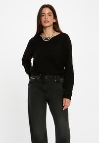 Maglione nero a maglia con scollo a barchetta e maniche lunghe, abbinato a jeans neri a vita alta con una leggera texture. Collana in catena d'argento come dettaglio.