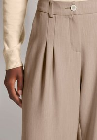 Lichtbeige broek met een geplooide voorkant, zijzak en een knoopsluiting. De stof heeft een soepele textuur met subtiele verticale lijnen.