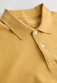Polo jaune moutarde pour homme avec col et deux boutons blancs, étiquette "NEXT Regular Fit M" à l'intérieur du col.