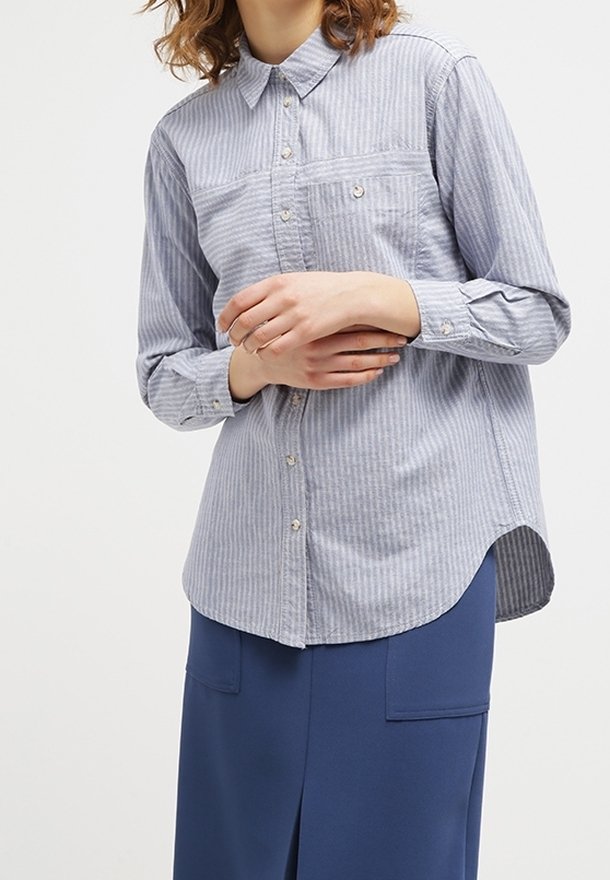 Personne portant une chemise à rayures bleu clair à manches longues et une jupe bleu marine avec des poches devant, les mains doucement jointes devant.