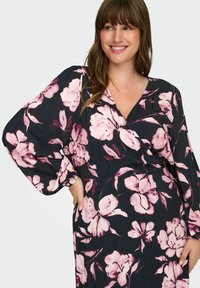 Žena se smije i nosi crnu haljinu s ružičastim cvjetnim uzorkom i dugim napuhanim rukavima, s jednom rukom na kuku, na svijetloj pozadini.