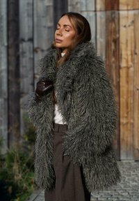 Kobieta z zamkniętymi oczami, ubrana w dużą szarą futrzaną kurtkę z puszystym kołnierzem, brązowe spodnie i jedną ciemną skórzaną rękawiczkę, stojąca na zewnątrz przy drewnianych ścianach.