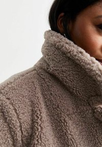 Giacca in poliestere fuzzy taupe con colletto alto, caratterizzata da una superficie testurizzata e dettagli di cucitura sottili.