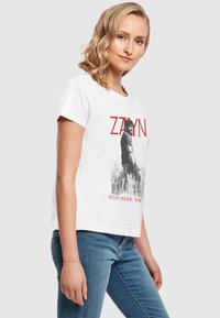 Hvid bomulds T-shirt med et grafisk print af en figur i sort mod højt græs, med rød tekst "ZAYN" og "ROOM UNDER THE".