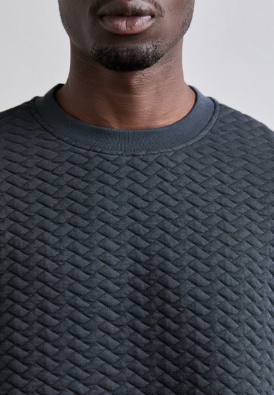 Sweatshirt gris foncé avec un motif matelassé texturé, un col rond et des poignets côtelés. Vue en gros plan mettant en avant les détails du matériau.