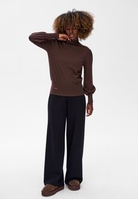 Kobieta z kręconymi włosami, ubrana w brązowy sweter z długim rękawem, czarne szerokie spodnie i brązowe buty, stojąca na tle jednolitego białego tła.