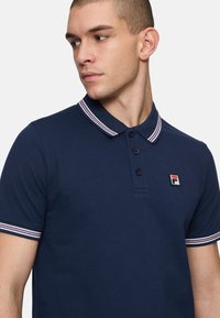 Marineblauw poloshirt met een kraag met rode en witte strepen, drie-knops knoopsluiting en een kleine FILA-logo patch op de borst.