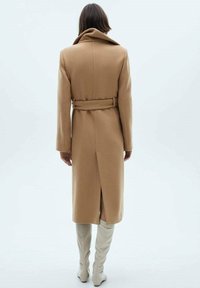 Kabát z velbloudí vlny se zavazovacím páskem v pase, velkým límcem a délkou do poloviny lýtek. Má zadní rozparek pro snadnější pohyb.