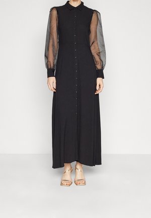 Černé maxi šaty s předním zapínáním na knoflíky, průsvitnými nabíranými rukávy a jednobarevnou sukní. Vycházejí s zlatými sandály na tenkých páscích.