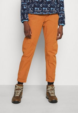 Persona con pantalones naranjas de pierna recta, botas de senderismo marrones con cordones beige y un suéter azul con patrón, de pie sobre un suelo gris.