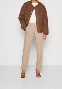 Brown quilted jacket featuring a round neckline and snap buttons, paired with loose beige pants and brown strappy heeled sandals.